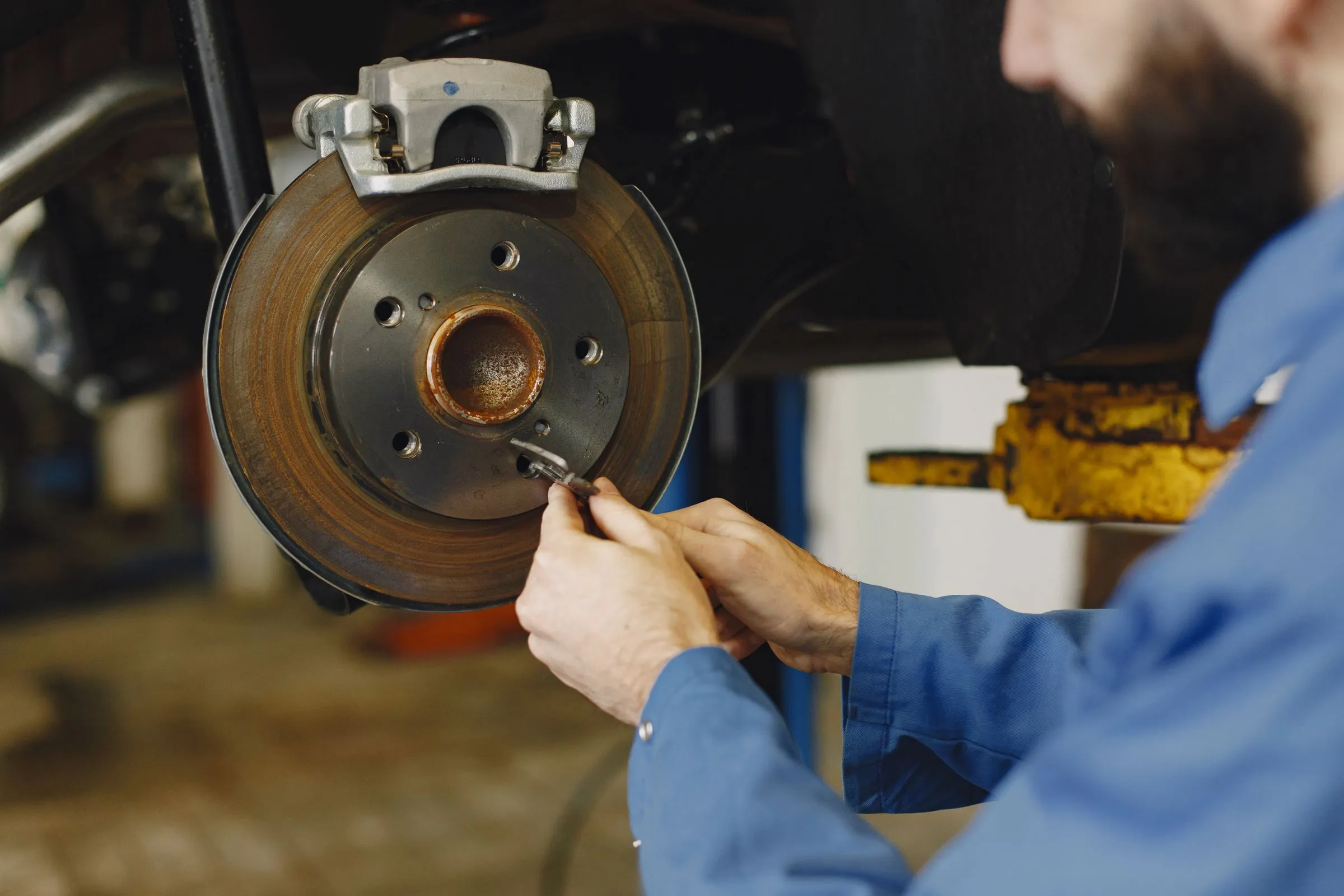 A mechanic in a blue work shirt measuring a worn brake rotor on a car raised on a lift.