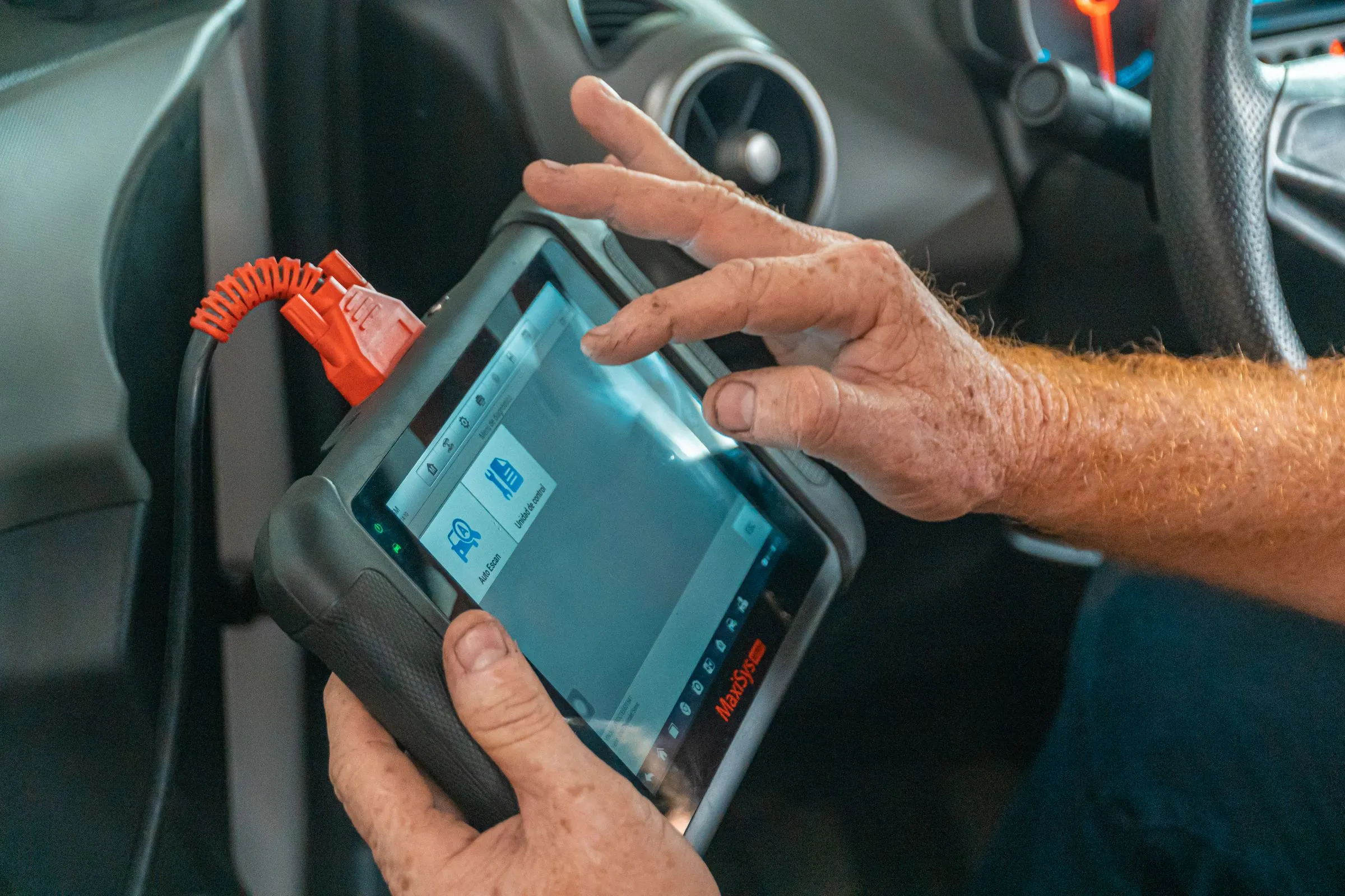 A technician’s hand on a diagnostic tablet plugged into a car’s port by a red cable.